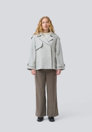Manteau en laine gris, surdimensionné, avec un design croisé, un large col et des poignets boutonnés, porté par-dessus un pull à col roulé beige et un pantalon marron.