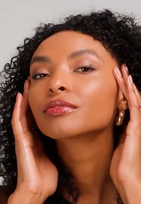 Femme aux cheveux bouclés touchant son visage, portant des boucles d'oreilles dorées en anneau et un rouge à lèvres rose brillant, sur un fond neutre.