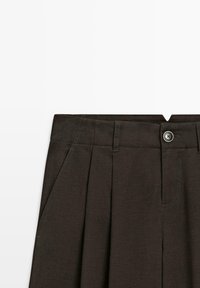 Jupe plissée marron foncé avec passants pour ceinture, poches latérales et fermeture par bouton argenté à la taille sur fond blanc.