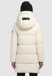 Longue doudoune crème avec capuche, design matelassé et un patch logo noir. Un bonnet en tricot noir côtelé est porté sur la tête.