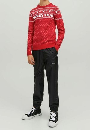 Personne portant un pull de Noël rouge avec le texte « MERRY XMAS », un pantalon de jogging noir et des baskets montantes noires, debout les mains dans les poches.