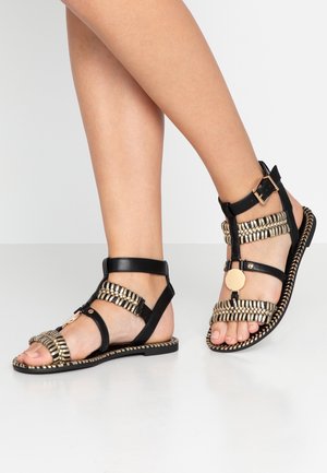 Czarna sandałka ze złotymi akcentami, z designem typu T-strap, płaską podeszwą, plecioną teksturą i okrągłą ozdobą na przodzie.