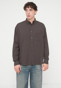 Πουκάμισο με κουμπιά σε σκούρο καφέ χρώμα με καρό σχέδιο. Διαθέτει γιακά, μακριά μανίκια και τσέπη στο στήθος. Φορεμένο με τζιν ανοιχτού μπλε.