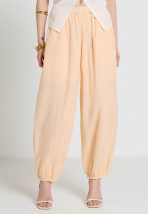 Femme portant un pantalon ample pêche avec des poignets élastiques, des sandales blanches, un haut blanc à manches courtes et des bracelets dorés sur un fond clair.