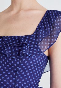 Vestido navy con lunares en azul claro; presenta un detalle de hombro con volantes transparentes y una tela texturizada, con un corsé ajustado y suaves pliegues.
