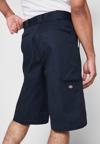 Homme portant un short bleu marine jusqu'aux genoux avec des poches à l'arrière et sur les côtés, ajustant la taille, debout devant un fond clair uni.