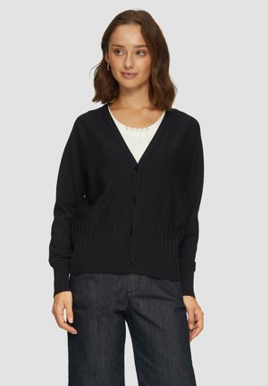 Schwarze Strickjacke, V-Ausschnitt-Design mit Knopfverschluss, gerippter Saum und Bündchen, weiche Textur, getragen über einem weißen Oberteil, kombiniert mit dunklen Hosen.