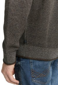 Gestructureerde bruine trui met contrasterende onderrand en manchetten, gedragen over blauwe spijkerbroek met omlijnd patroon op de achterzakken.