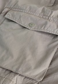 Zbliżenie na beżową kieszeń z tkaniny z patką zabezpieczoną pojedynczym okrągłym zatrzaskiem na ubraniu.