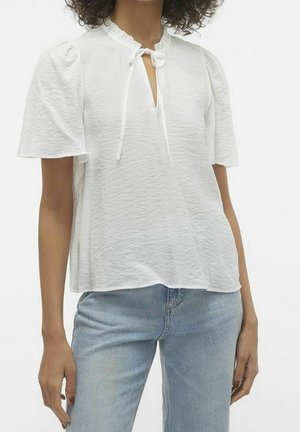 Femme portant un chemisier blanc texturé à manches courtes avec un nœud volanté au col, associé à un jean en denim bleu clair.