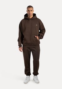 Man som står klädd i en mörkbrun hoodie och matchande joggers med vita sneakers, poserar mot en enkel ljus bakgrund.