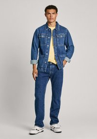Chaqueta de mezclilla con dos bolsillos en el pecho, cierre de botones y puños enrollados, combinada con jeans a juego y una camiseta amarilla, llevada con zapatillas blancas.