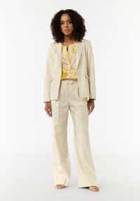 Beige maßgeschneiderter Anzug mit einem strukturierten Blazer und weit geschnittenen Hosen. Darunter ein leuchtend gelbes Oberteil mit einem auffälligen Blattmuster.