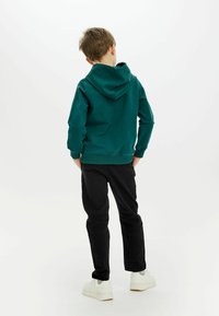Grön huvtröja i bomull med ribbade muddar och nederkant, tillsammans med svarta byxor och vita sneakers, visad bakifrån.