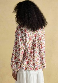 Biała bluzka w kwiaty z czerwonymi i niebieskimi motywami, długie rękawy z elastycznymi mankietami, falbany oraz luźny krój. Pokazany widok z tyłu.