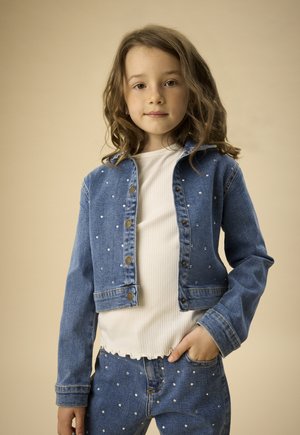 Jong meisje met golvend bruin haar, gekleed in een spijkerjasje met studs en een spijkerbroek, met een wit geribd shirt, staand tegen een beige achtergrond.