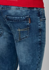 Donkerblauwe denim jeans met een achterzak met gele stiksels en een klein bruin logo-label, die een licht versleten textuur tentoonstelt.