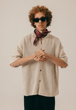 Personne aux cheveux bouclés, portant des lunettes de soleil noires, un foulard bordeaux autour du cou, une chemise beige à boutons perforée, et un tatouage sur l'avant-bras, mains jointes.