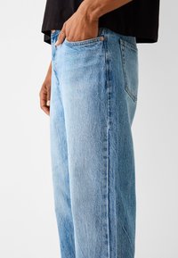 Jasnoniebieskie dżinsy z denimu o luźnym kroju, widocznym przeszyciu kieszeni oraz subtelnej fakturze ziarnistej. Posiadają wysoki stan i zwężane nogawki.