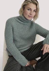 Hellgrauer, gestrickter Rollkragenpullover mit gerippter Textur, lockerer Passform und langen Ärmeln, kombiniert mit Cordhosen.