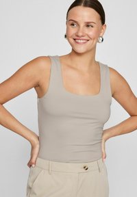 Beige sleeveless bodysuit with a square neckline, made of smooth, stretchy fabric, paired with light beige trousers and round earrings.
