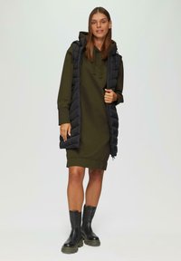 Een olijfgroen hoodie jurkje tot de knie onder een zwarte puffer vest. De vest heeft een ritssluiting en een gestikte textuur. Zwarte enkellaarzen met dikke zolen.