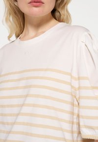 Camiseta blanca con rayas verticales beige, con cuello redondo y corte holgado. Detalle bordado sutil en el pecho. Material de algodón suave.