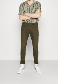 Jeans ajustados de color verde oliva con una textura suave, combinados con una camisa de manga corta de estampado. Completados con zapatillas blancas con detalles negros.