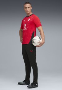 Rød fodboldtrøje med tekst og mønsterdetaljer, sorte træningsbukser og sorte støvler. Model holder en hvid fodbold med sorte accenter.