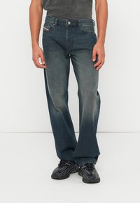 Muž v vybledlých tmavě modrých džínech s rovným střihem a černých sportovních botách, stojící před jednobarevným bílým pozadím.