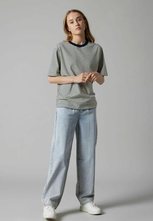 Junge Person steht mit verschränkten Händen, trägt ein schwarz-weiß gestreiftes T-Shirt, hellblaue Hose mit weitem Bein und weiße Turnschuhe.