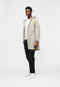 Νεαρός άντρας στέκεται φορώντας ανοιχτό μπεζ μπουφάν με κουκούλα πάνω από λευκή μπλούζα, μαύρο παντελόνι και λευκά αθλητικά παπούτσια, μπροστά σε μονόχρωμο φόντο.