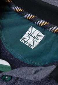 Donkere stoffen kraag met groene binnenkant, voorzien van een label met een Union Jack, "J&JOY," en tekst. Geaccentueerd met een gestreept patroon in geel en wit.