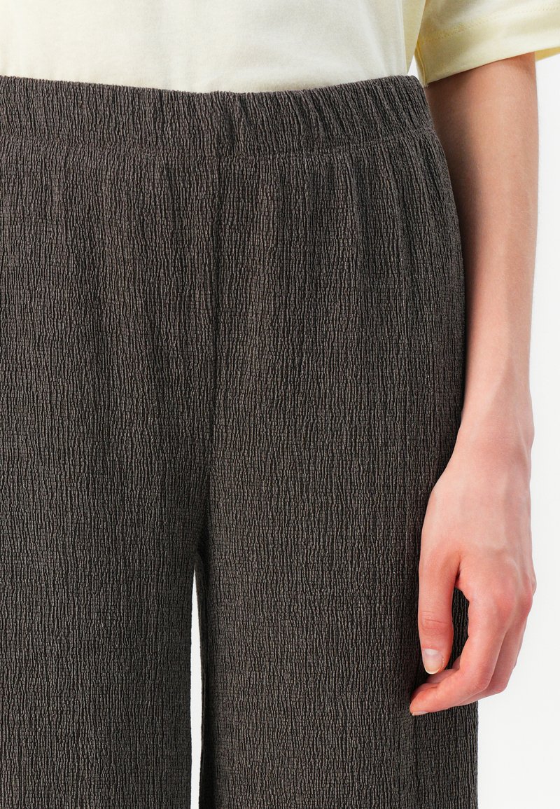 Donkerbruin textuurpantalon met een gekreukt materiaal. De tailleband is elastisch en zorgt voor een geplooide uitstraling. Hand gepositioneerd bij de zijkant.