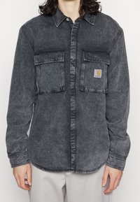 Tmavě šedá denimová košile s knoflíkovou přední částí, dvěma náprsními kapsami a nášivkou loga Carhartt. Obsahuje klasický límec a dlouhé rukávy.