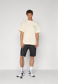 Beige bomull T-skjorte med et grønt grafisk motiv på brystet, kombinert med svarte shorts. Hvite knelengde sokker og svarte treningssko fullfører antrekket.