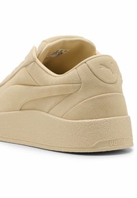 Zapatilla de ante beige con una suela de goma texturizada, forma redondeada y un branding mínimo, que presenta un logotipo sutilmente grabado en el talón.