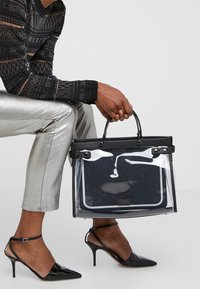 Borsa tote trasparente con rifiniture nere e manici, design strutturato, abbinata a pantaloni metallici argentati e tacchi alti neri.