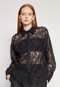 Svart blondebluse med knapp foran, med intrikate blomstermønstre og en gjennomsiktig design. Lange ermer med klassisk krage.
