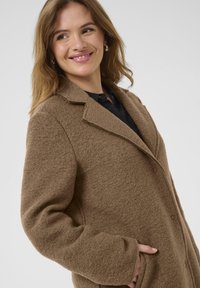 Cappotto marrone, texturizzato, con revers a punte, chiusura con bottoni e tasche laterali. Tessuto morbido e vestibilità rilassata.