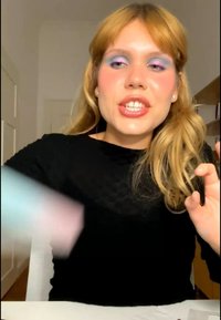 Femme aux cheveux blonds et maquillage coloré aux yeux parle et fait des gestes de la main droite assise à une table à l'intérieur.