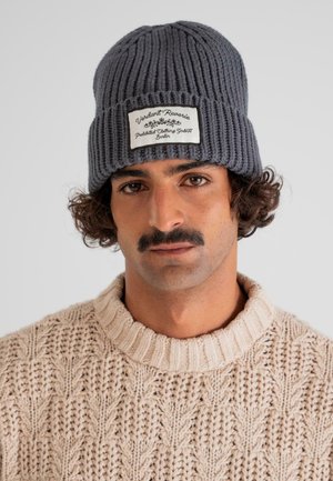 Man with curly hair and mustache wearing a gray knit beanie with a label and a cream cable-knit sweater against a neutral background.