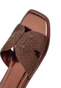 Sandale plate marron avec un design de lanières tressées, bout carré et accents métalliques brillants sur la semelle intérieure. Texture lisse dans l'ensemble.