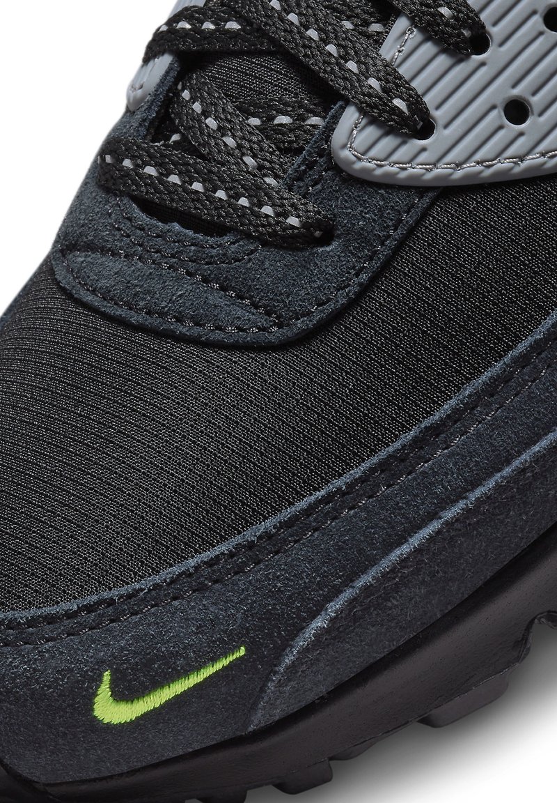 Zapatilla Nike con una parte superior de malla negra, acentos de ante texturizado, un logo verde y una suela de goma negra duradera. Los cordones son de color gris oscuro.