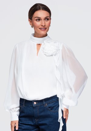 Femme portant un chemisier blanc transparent à manches longues avec un détail floral sur l'épaule et un jean bleu foncé, regardant sur le côté.