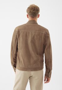 Hnědá semišová bunda s volným střihem, s kontrastním šitím, uzavřeným límcem a detaily na zapínání rukávů. Zadní pohled na jednoduchém pozadí.