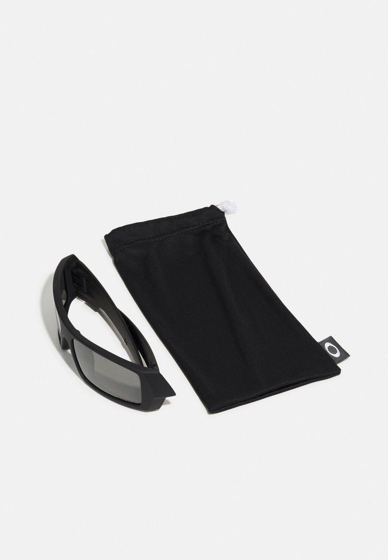 Gafas de sol negras con lentes oscuros, diseño de marco angular; acompañadas de un suave estuche negro con un cordón blanco y etiqueta de logo.