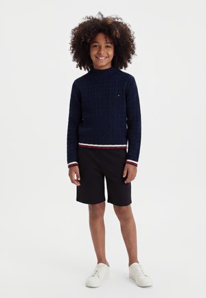 Enfant aux cheveux bouclés portant un pull bleu marine, un short noir et des baskets blanches, debout et souriant sur un fond blanc.
