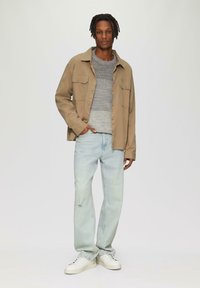 Veste boutonnée tan sur un pull gris, jeans bleu clair avec une légère déchirure, et baskets blanches. Le modèle se tient devant un fond neutre.