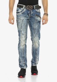 Schlanke, hellblaue Jeans mit Distressed-Details, mehreren Reißverschluss-Taschen und kontrastierendem Nähen; getragen zu schwarzen, mit Nieten besetzten Turnschuhen.